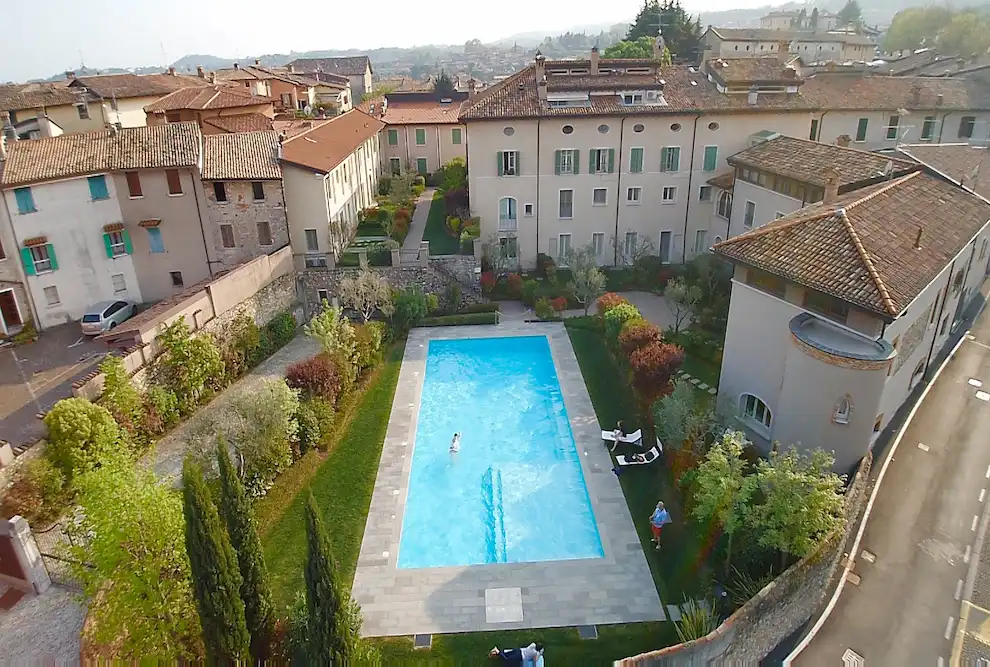 Ripresa aerea della piscina a sfioro e del giardino di Palazzo Scolari a Polpenazze, con vista sui tetti del borgo storico e sulle colline del Garda.